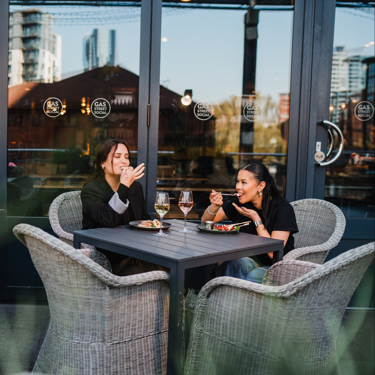 Two women sitting at a outdoor terrace dining table with wicker chairs inside a restaurant, with large glass windows reflecting the city skyline behind them.