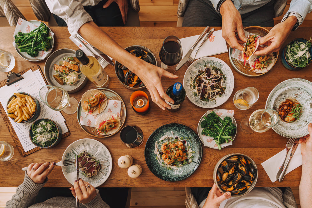 Overhead view of a wooden dining table filled with plates of food, including pasta, greens, fries, mussels, and drinks, with multiple people's hands and arms visible sharing a meal.