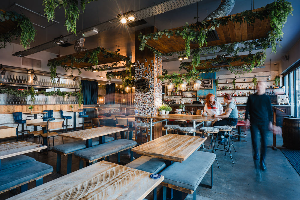 Spacious Gas Street Social restaurant interior at The Mailbox in Birmingham, featuring communal wooden tables, blue seating, hanging greenery and a stylish cocktail bar.